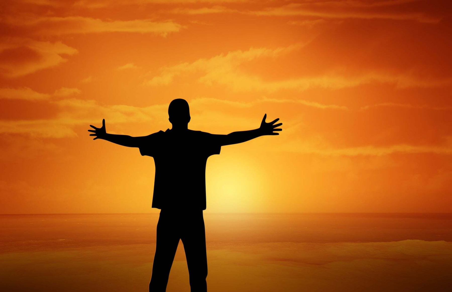Auto-generated description: A silhouette of a person with outstretched arms stands against a vibrant orange sunset over a calm body of water.