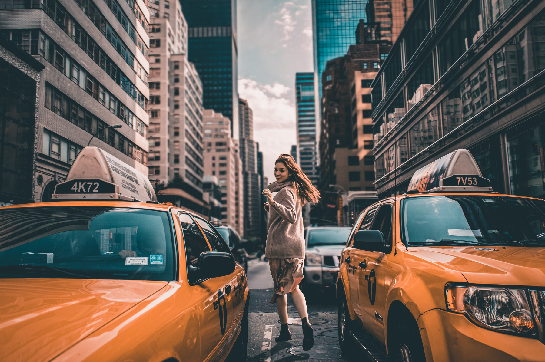 A woman is walking between two yellow taxis on a busy city street lined with tall buildings.